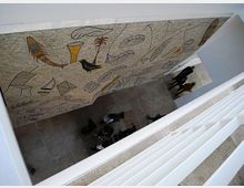 Vista dall’alto di un atrio interno di edificio moderno con pavimento in pietra e pareti bianche, attraversato da una passerella. Su una grande parete laterale è presente un ampio murale astratto con simboli e forme stilizzate in colori ocra e nero.