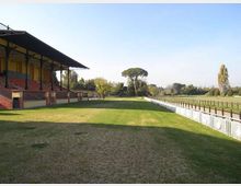 Vista di un campo erboso lungo una tribuna coperta con struttura in metallo e gradinate in muratura. A destra corre una lunga recinzione bianca che delimita un’area aperta, con alberi e vegetazione sullo sfondo sotto cielo sereno.