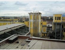 Vista su un complesso di edifici industriali gialli con tetti piani, passerelle coperte e condotte verticali in metallo su una torre centrale. Sullo sfondo si vedono palazzi residenziali e un cielo nuvoloso.