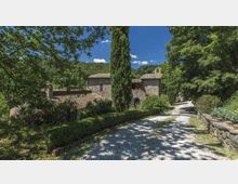 Viale sterrato di ghiaia bordato da siepi e alberi alti, che conduce a un complesso di edifici in pietra con tetti in coppi. Sullo sfondo si vedono colline boscose sotto un cielo azzurro.