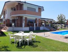 Villa a due piani con facciata in mattoni e intonaco bianco, terrazzi con ringhiere metalliche e finestre con grate decorative. In primo piano giardino con tavolo e sedie bianche su prato, vialetto in mattoni e piscina rettangolare sul lato destro.