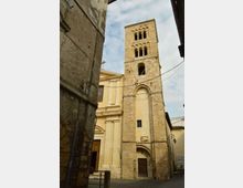 Campanile romanico in pietra con finestre ad arco sovrapposte e ingresso ad arco alla base, affiancato da una chiesa intonacata color chiaro. La torre si affaccia su una stretta piazza tra edifici in muratura, con cavi sospesi e cielo parzialmente nuvoloso sullo sfondo.