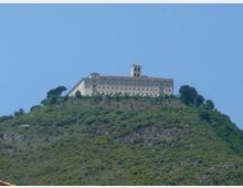 Grande edificio in muratura chiara, simile a un monastero o santuario, posto sulla sommità di una collina verde con terrazze e vegetazione. Il complesso ha una lunga facciata con file di finestre e una torre campanaria centrale, circondato da pochi alberi sotto un cielo azzurro.