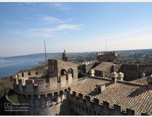 Veduta dall’alto di un castello in pietra con mura merlate, torri e tetti in coppi, con cortile interno e arcate. Sullo sfondo si estendono un lago e colline rurali sotto un cielo sereno.