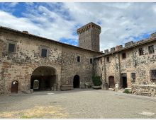 Cortile interno di un castello in pietra, circondato da edifici con muri in tufo e mattoni, finestre piccole e portali ad arco. Sullo sfondo si vede una torre quadrata merlata e un camminamento con merli sotto un cielo parzialmente nuvoloso.