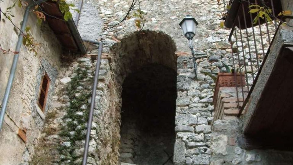 Cortile stretto in un borgo con edifici in pietra, un grande arco che conduce a una scalinata e muri con finestre e persiane verdi. In primo piano si vedono roccia e vegetazione, con rami di albero sopra e un lampione fissato alla parete.