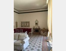 Salotto in stile tradizionale con divano chiaro, pareti bianche con decori e cornici, e pavimento a piastrelle con motivo geometrico e bordura. Ampie tende chiare filtrano la luce su arredi d’epoca, tra cui sedie in legno, un camino e un tavolino.