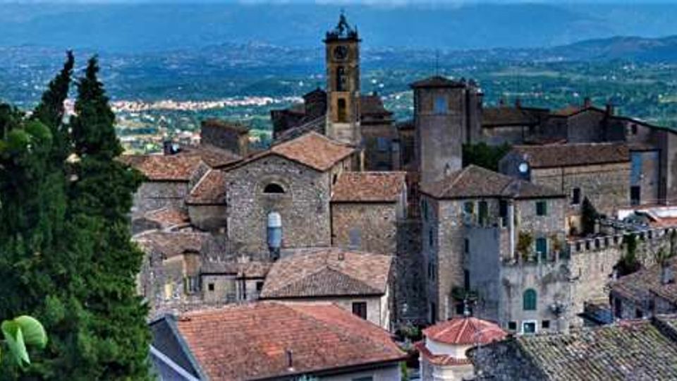 Veduta panoramica di un borgo storico su una collina, con case in pietra e tetti in coppi e un campanile che svetta tra gli edifici. Sullo sfondo si estende una valle con campi e insediamenti, incorniciata da colline e montagne lontane.