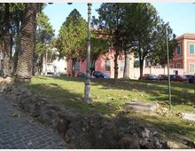 Piccolo parco urbano con prato in lieve pendenza, alberi alti e un lampione centrale, bordato da un muretto di pietra e da una pavimentazione in ciottoli. Sullo sfondo si vedono edifici con facciate rosa e bianche e alcune auto parcheggiate lungo la strada.