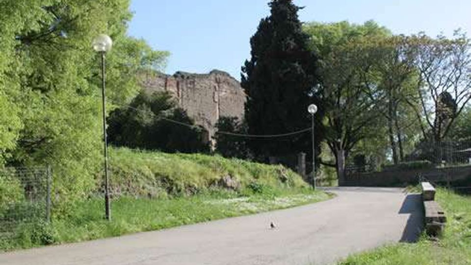 Viale asfaltato in un parco con prato e alberi ai lati, illuminato da lampioni. Sullo sfondo si vedono resti di mura in pietra su una collina e un alto cipresso vicino a un cancello.