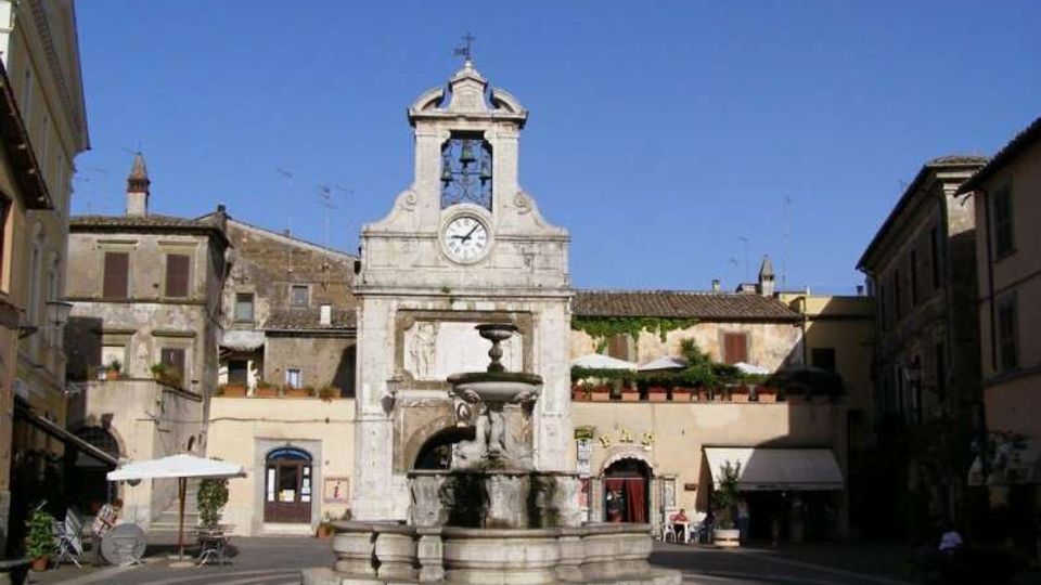 Piazza storica con una fontana in pietra al centro e, sullo sfondo, una facciata monumentale con campanile e orologio. Intorno si vedono edifici antichi a più piani con finestre e persiane, sotto un cielo sereno.