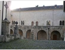 Cortile in pietra con pavimentazione in ciottoli, fiancheggiato da un edificio storico in muratura chiara con archi e finestre ad arco. In primo piano si vedono una croce in ferro su piedistallo e muretti in pietra, con un passaggio ad arcate al livello inferiore.