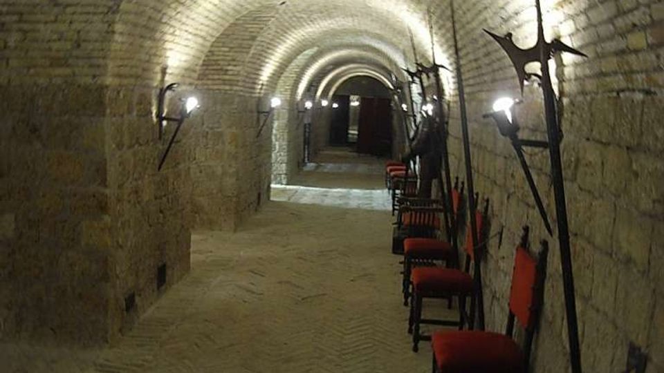 Corridoio sotterraneo in pietra con volta a botte in mattoni, illuminato da lampade a parete lungo entrambi i lati. Sul lato destro sono allineate sedie con seduta rossa e sono appoggiate lunghe armi ad asta contro il muro, mentre il passaggio prosegue fino a una porta in fondo.