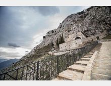 Scalinata in pietra con ringhiera metallica che sale lungo un pendio roccioso verso un edificio storico in pietra addossato alla montagna. Sullo sfondo si vedono pareti di roccia scoscesa, vegetazione rada e cielo nuvoloso.