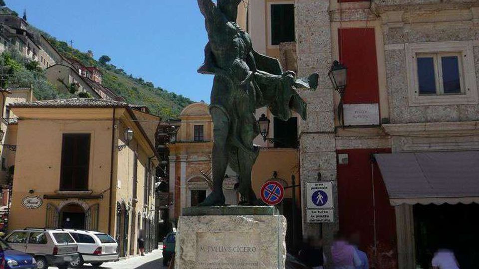 Statua in bronzo su alto basamento in pietra al centro di una piccola piazza, circondata da edifici storici con facciate colorate e balconi. Sullo sfondo si vede una strada con auto parcheggiate e una collina verde sotto cielo sereno.