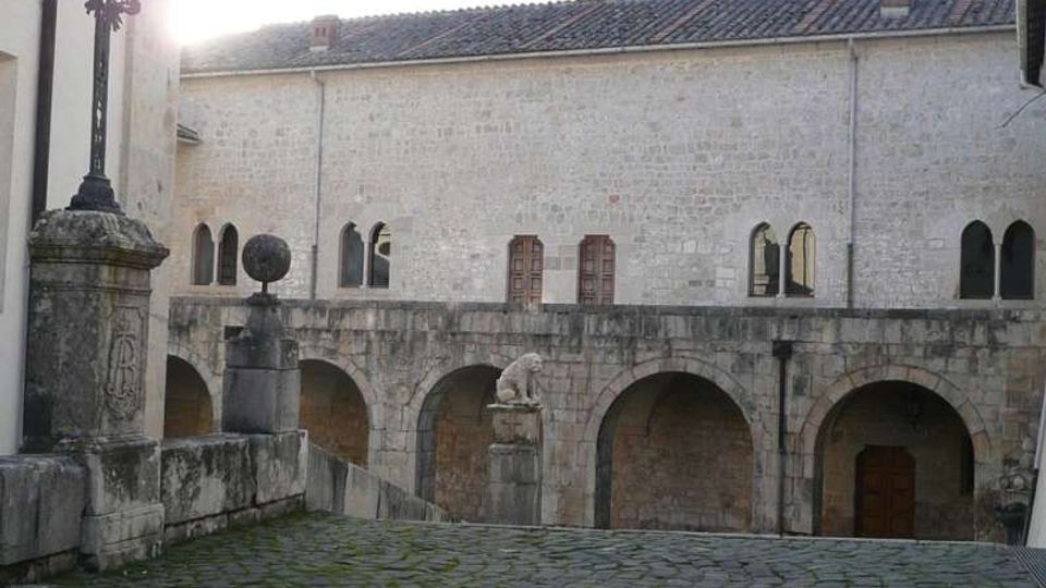 Cortile in pietra con pavimentazione in ciottoli, fiancheggiato da un edificio storico in muratura chiara con archi e finestre ad arco. In primo piano si vedono una croce in ferro su piedistallo e muretti in pietra, con un passaggio ad arcate al livello inferiore.