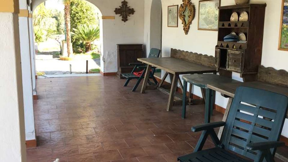 Veranda coperta in stile rustico con pavimento in cotto, soffitto con travi in legno scuro e pareti bianche decorate con piatti e cornici. Lungo la parete ci sono tavoli e sedie in plastica e un mobile credenza; un grande arco aperto conduce al giardino esterno.