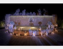 Fontana semicircolare in una piazza lastricata, illuminata di notte e circondata da numerose lanterne e candele. Sullo sfondo c’è un alto muro in pietra con un simbolo circolare e rocce decorative in sommità.