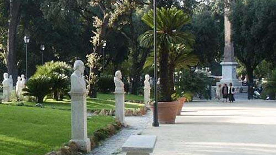 Viale pedonale in un parco con ampi prati e alberi alti, fiancheggiato da statue e busti in pietra su piedistalli. Lungo il percorso si vedono lampioni, una panchina, grandi vasi con palme e un alto monumento/obelisco sullo sfondo.