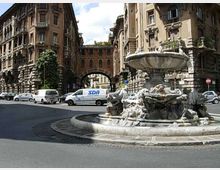Rotonda urbana con grande fontana in pietra scolpita al centro, circondata da strada asfaltata. Sullo sfondo si vedono palazzi storici in stile monumentale con facciate in muratura, balconi e un arco che collega due edifici, con auto parcheggiate lungo i lati.