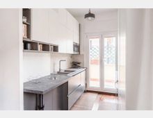 Cucina moderna e stretta in stile lineare, con mobili bianchi e grigi, piano di lavoro in pietra, lavello e piano cottura integrati e scaffali a vista. In fondo, una portafinestra a due ante illumina l’ambiente e si affaccia su un balcone con griglia decorativa.