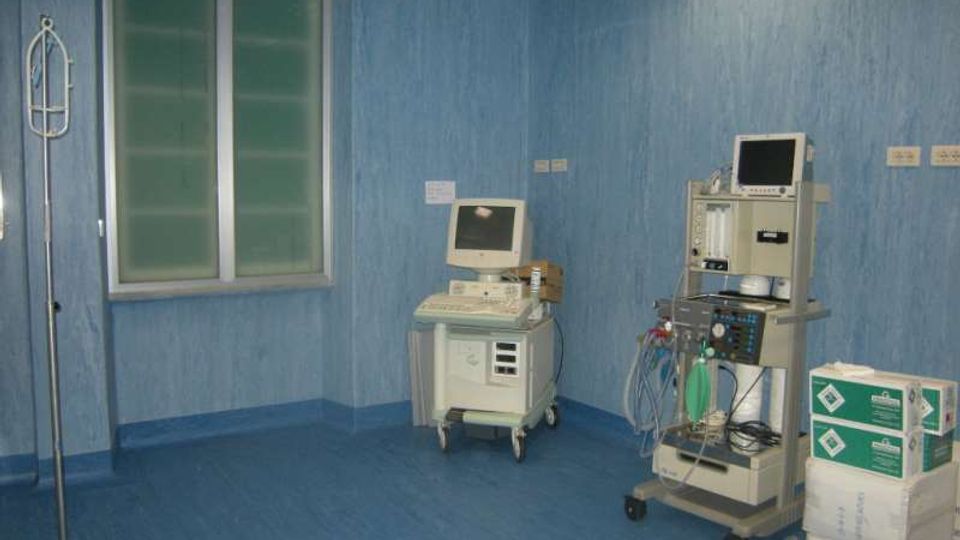 Stanza clinica con pareti e pavimento blu, finestra a due ante e attrezzature mediche su carrelli, tra cui un monitor e una macchina per anestesia o ventilazione con tubi e bombola. A lato si vedono un supporto per flebo e scatole di forniture impilate.