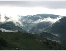 Panorama di una valle montana con colline e montagne verdi, attraversata da banchi di nebbia e nuvole basse. In primo piano si vedono pendii erbosi con pochi alberi e alcune case sparse.