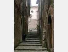 Vicolo stretto in un centro storico, con una lunga scalinata in pietra e pavimentazione a ciottoli tra alti edifici in muratura. In fondo si vede una facciata chiara con finestra rotonda e un lampione a parete lungo il passaggio.