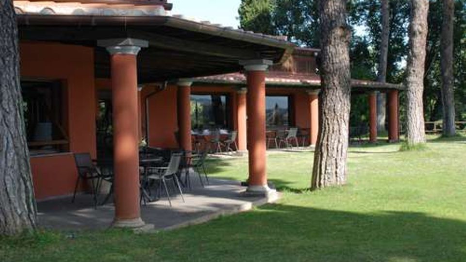 Veranda coperta di una struttura in stile rustico con colonne cilindriche color terracotta e tetto in tegole, arredata con tavoli e sedie. L’edificio è circondato da un prato verde con alti pini e alberi sullo sfondo.