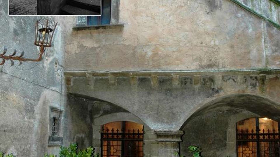 Cortile interno di un edificio storico in pietra e intonaco, con scala esterna in muratura che sale lungo la parete e un porticato ad archi con colonne. Sul pavimento in mattoni a spina di pesce ci sono grandi vasi con piante, e sullo sfondo si vedono porte con grate in ferro battuto.
