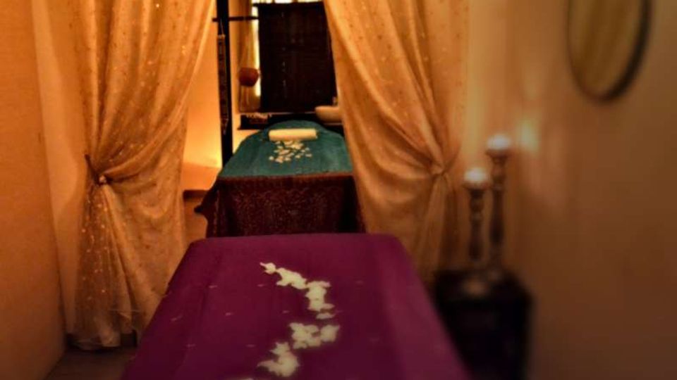 Interno di una spa o sala massaggi con due lettini allineati, coperti da teli viola e turchese decorati con petali bianchi. Tende leggere color crema dividono lo spazio e sullo sfondo si vede una finestra con tende scure e luci soffuse.