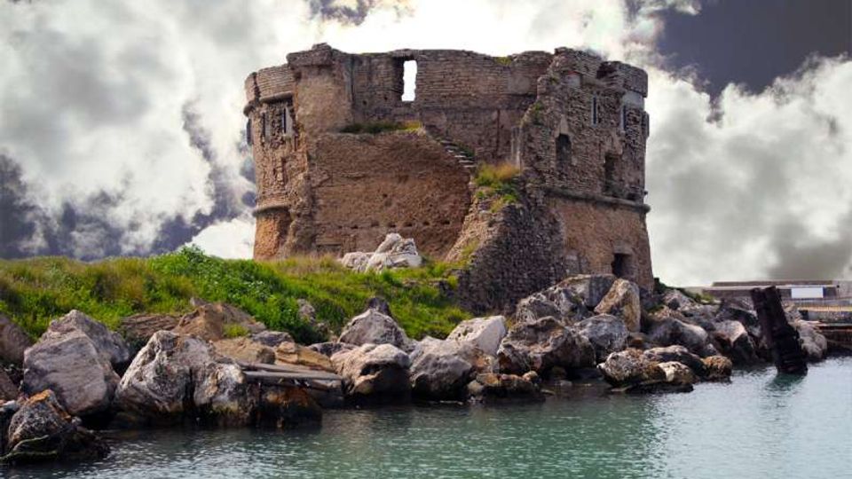 Rovine di una torre in pietra su un piccolo promontorio erboso, circondata da massi lungo la riva. In primo piano si vede l’acqua calma e sullo sfondo un cielo nuvoloso.