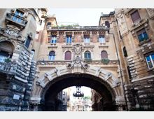 Arco monumentale in pietra che collega due edifici storici, con facciata decorata da bassorilievi e stemmi scolpiti al centro. Sotto la grande volta si intravede una strada urbana e un lampadario sospeso.
