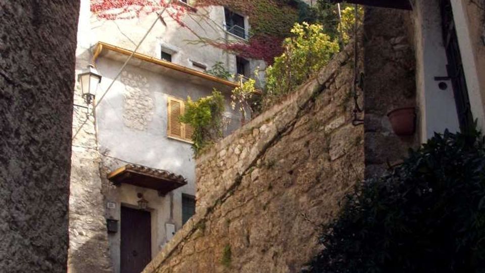 Vicolo stretto in un borgo storico, con scalini in pietra e un alto muro in muratura che sale verso case in intonaco e pietra. Si vedono persiane, un lampione a parete, tetti sporgenti e cavi sospesi tra gli edifici.