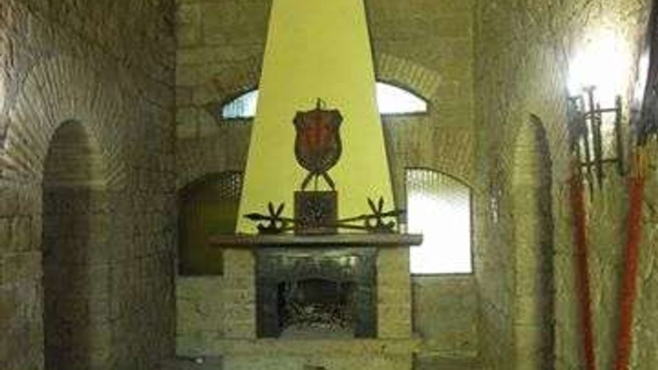 Interno di una sala in pietra con soffitto a volta e pavimento in mattoni disposti a spina di pesce. Al centro c’è un grande camino con cappa alta e chiara, sormontato da uno stemma, con finestre ad arco e torce o supporti lungo le pareti laterali.