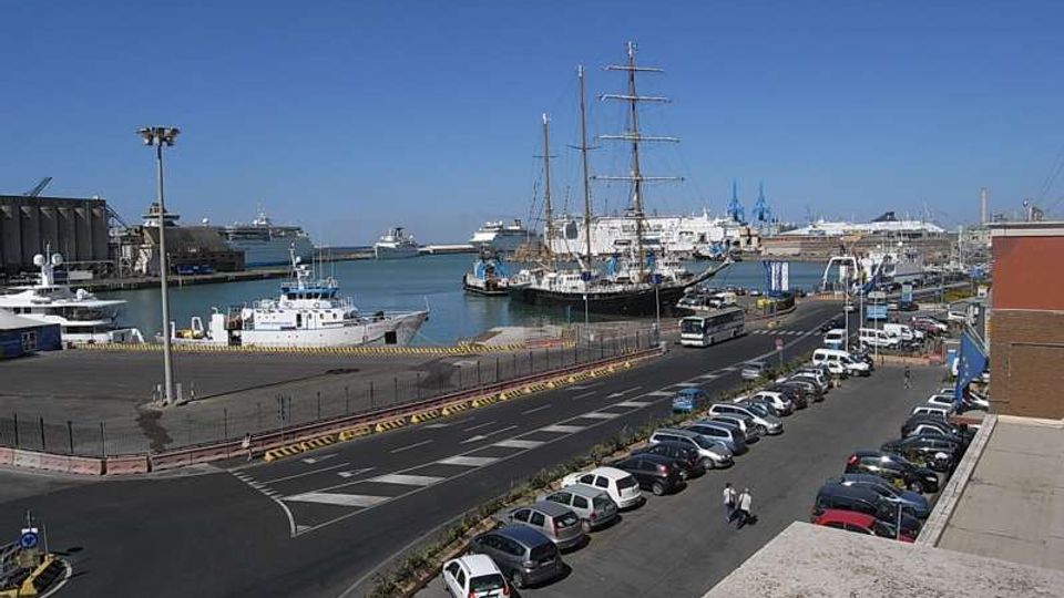 Veduta di un porto con banchine e parcheggi, una strada a più corsie in primo piano e diverse imbarcazioni ormeggiate, tra cui un grande veliero con alberi alti. Sullo sfondo si vedono navi più grandi e gru portuali sotto un cielo sereno.