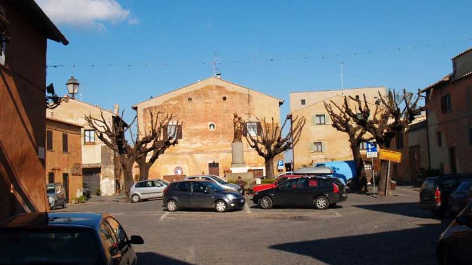 Piazza di un piccolo centro con parcheggio asfaltato e auto in sosta, circondata da edifici in muratura dai toni ocra. Al centro si vede una statua su piedistallo e una fila di alberi potati senza foglie sotto un cielo sereno.