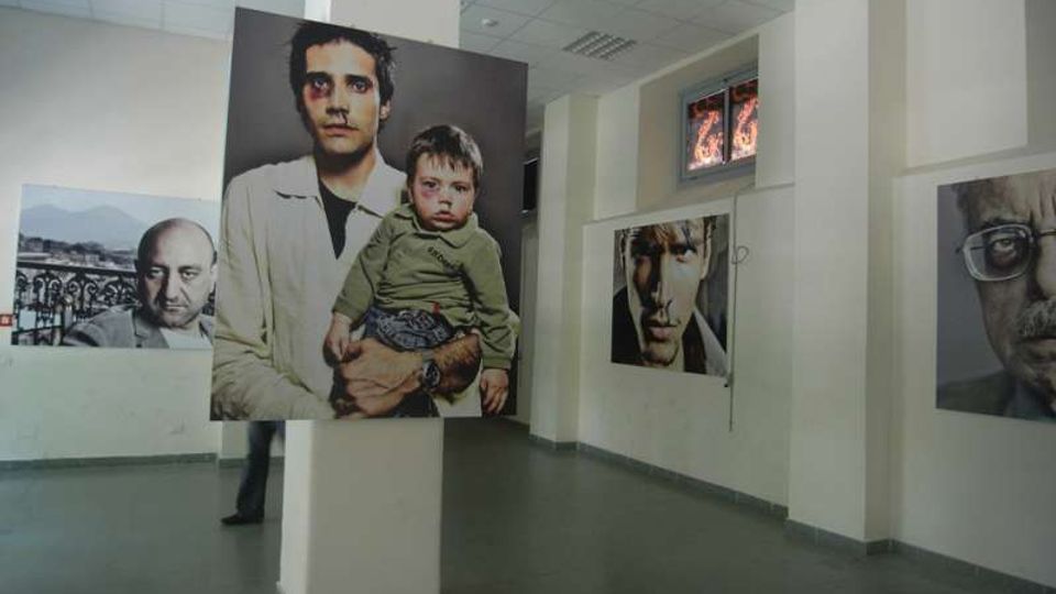 Interno di una galleria o spazio espositivo con pavimento grigio e soffitto a pannelli, scandito da pilastri bianchi. Sulle pareti e su un pilastro sono appesi grandi ritratti fotografici, con una finestra in alto sul fondo.