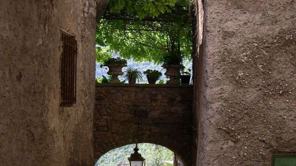 Passaggio stretto in pietra con grandi archi sovrapposti, che incorniciano una terrazza con ringhiera e piante in vaso sotto una pergola verde. In fondo si vede una discesa tra muri antichi con un lampione sospeso e vegetazione sullo sfondo.