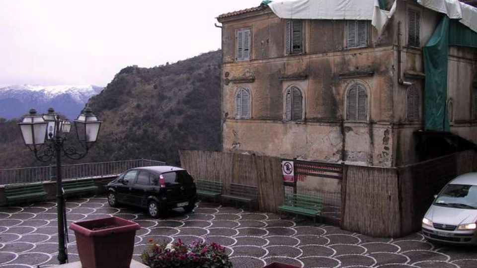 Piazzale panoramico pavimentato con motivi circolari bianchi e neri, con panchine, lampioni e alcune auto parcheggiate. Sul lato destro si vede un edificio in muratura vecchio e deteriorato con impalcatura e teli, sullo sfondo colline e montagne innevate.