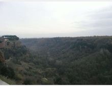 Ampia valle boscosa vista dall’alto, con pareti rocciose e un altopiano sullo sfondo sotto un cielo nuvoloso. Sul bordo della scarpata a sinistra si intravedono edifici e un gruppo di alberi.