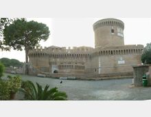 Veduta esterna di una grande fortificazione in mattoni con mura merlate e un’alta torre cilindrica, affacciata su una piazza lastricata. Intorno si vedono alberi ad alto fusto e vegetazione, con un basso muro di contenimento lungo il perimetro.