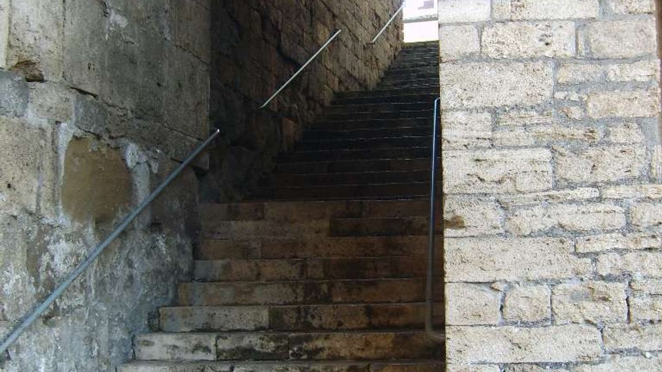 Scalinata in pietra consumata che sale all’interno di un passaggio ad arco, tra alti muri di muratura grezza. Corrimani metallici corrono lungo i lati e in cima si intravede luce naturale dall’apertura superiore.