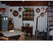 Interno rustico di una casa con soffitto a travi in legno, tavolo da pranzo con tovaglia e sedie, e pareti decorate con pentole di rame appese e una credenza con piatti e bottiglie. A destra si vede una scala a chiocciola metallica, con anfore e altri oggetti tradizionali disposti lungo le pareti.