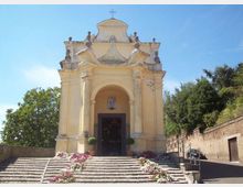 Facciata di una piccola chiesa in stile barocco, color crema, con portico ad arco, colonne e croce in sommità. Davanti, una scalinata in pietra con aiuole fiorite conduce all’ingresso, circondato da alberi e un muro laterale.