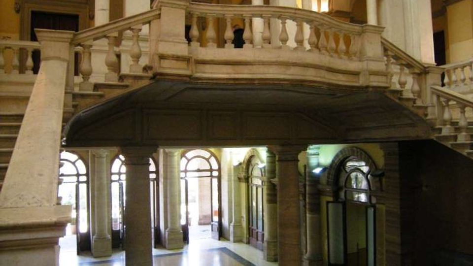 Interno di un edificio storico con grande scala monumentale e ballatoio in pietra con balaustra, sostenuti da colonne. Al livello inferiore si vedono archi e alte finestre ad arco che illuminano un ampio atrio.
