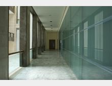 Corridoio interno di un edificio moderno con pavimento in marmo chiaro, colonne in pietra e grandi finestre sul lato sinistro. A destra una lunga parete vetrata con pannelli e telai metallici, con porta in fondo al corridoio.