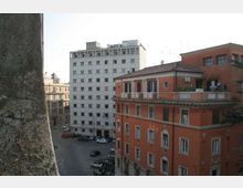 Veduta urbana con palazzo moderno bianco a più piani e edificio storico in mattoni rossi con terrazza e balconate; in basso si vede un incrocio stradale con auto parcheggiate tra altri edifici.