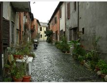 Vicolo stretto in un borgo con pavimentazione in ciottoli, fiancheggiato da case a due piani con facciate intonacate e persiane in legno. Lungo i muri ci sono numerosi vasi e fioriere con piante, e in fondo si intravede un edificio chiaro con tetto in coppi.