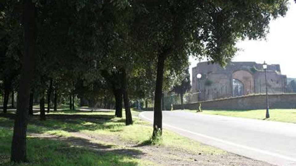 Viale alberato con ampie aree erbose e una strada asfaltata che curva verso destra. Sullo sfondo si vede una struttura storica in muratura con grandi archi, oltre un muretto e alcuni lampioni.